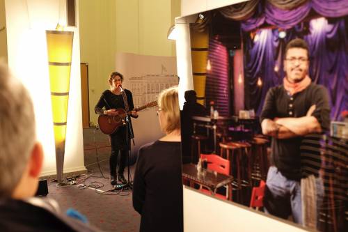 Eine Musikerin sing und spielt Gitarre in der Wandelhalle. Menschen hören ihr zu. Im Vordergrund sieht man ein Bild einer Ausstellung.
