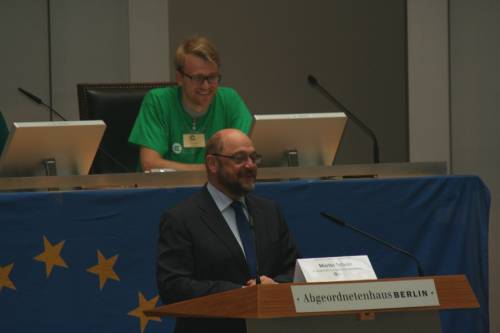 Martin Schulz steht am Rednerpult im Plenarsaal und lacht. Hinter ihm hängt eine europäische Flagge. Dahinter sitzt ein Mann im grünen T-Shirt und lacht ebenfalls.