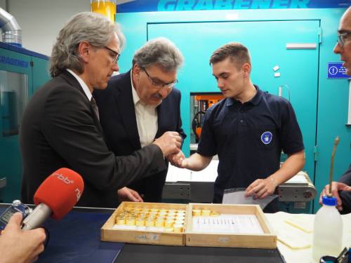 Ein junger Mann zeigt Ralf Wieland und einem anderen Mann eine Münze. Sie befinden sich in einer Produktionsstätte.