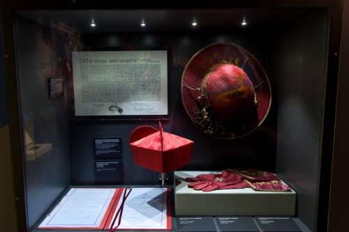 Diverse Ausstellungsexponate in einem gläsernen Kasten, u.a. zwei rote Hüte und rote Handschuhe.