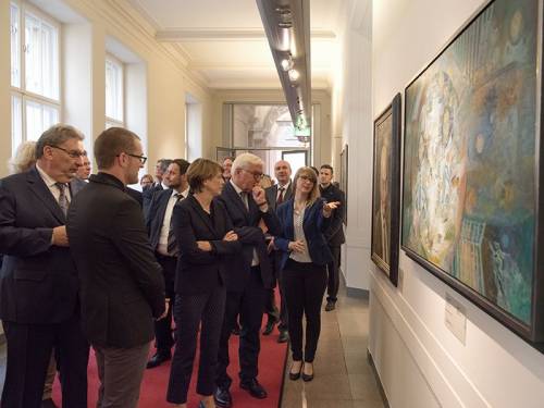 Frank-Walter Steinmeier, Ralf Wieland und weitere Personen sehen sich eine Ausstellung an. Eine Frau erklärt ein Gemälde.