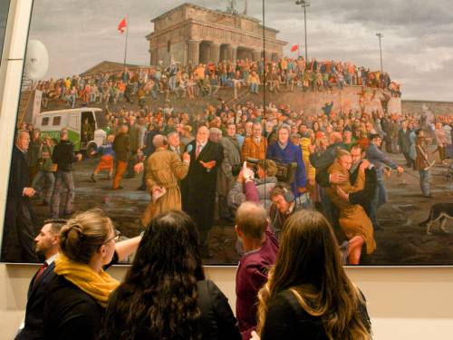 Männer und Frauen schauen sich ein großes Gemälde an, welches den Mauerfall in Berlin zeigt.