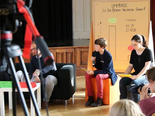 Jugendliche, eine mit mikrofon, sitzen auf Hockern bei einer Veranstaltung.