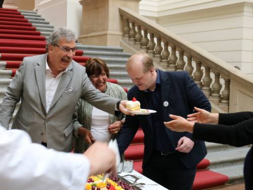Ralf Wieland reicht jemanden einen Teller mit Kuchen. Neben ihm stehen noch eine Frau und ein Mann, der sich zu einem Kuchenbüffett beugt. Sie sind in der Eingangshalle des Abgeordnetenhaus. Alle lachen.