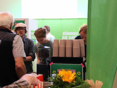 Ein Messestand von der Grünen Fraktion. Man sieht einen Kaffeeautomaten und Menschen die darum stehen.