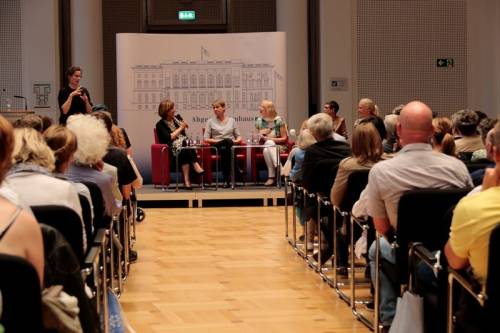 Vier Frauen diskutieren auf einer Podiumsdiskussion. Eine weitere Frau übersetzt in Gebärdensprache. Man sieht Publikum, welches zuhört.