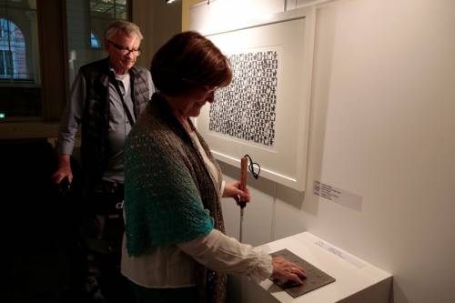 Besucher schauen sich eine Ausstellung im Abgeordnetenhaus Berlin an. Eine blinde Frau ertastet die Beschreibung zu einem Werk.