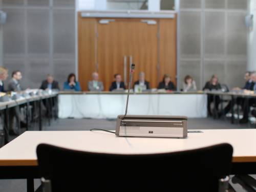 Ein leerer Sitz mit Mikrofon während einer Sitzung des Petitionsausschusses, im Hintergrund sieht man verschwommen andere Ausschussmitglieder