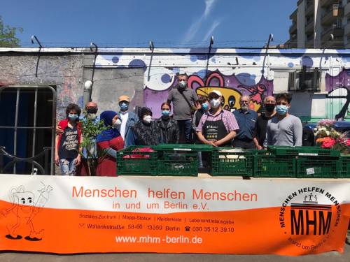 Menschen stehen hinter grünen Kästen und vor einer Wand mit Graffiti und Stacheldraht. Sie tragen Masken. Vor ihnen ein Plakat mit den Worten "Menschen helfen Menschen in und um Berlin e.V."