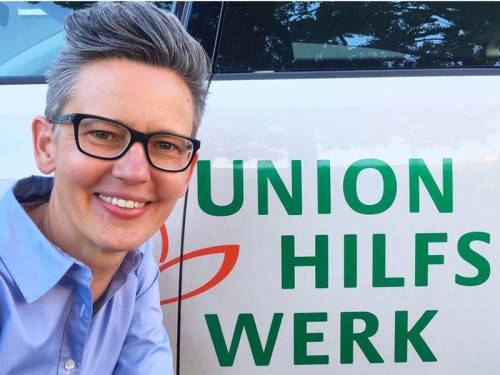 Eine Frau mit kurzen leicht grauen Haaren und Brille lacht in die Kamera. Sie hockt vor einem Auto mit der Beschriftung "Unionhilfswerk"