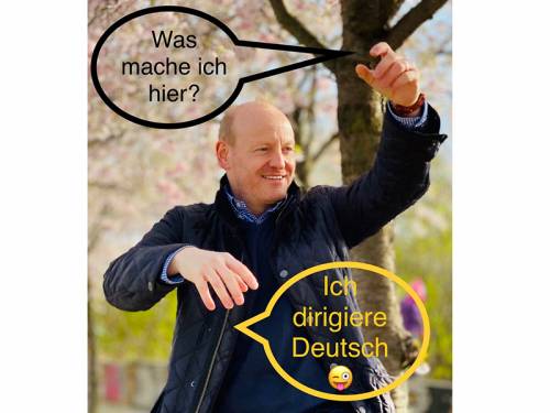 Ein Mann steht im Park und hebt die Arme, wie ein Dirigent. Zwei Sprechblasen beinhalten die Texte: "Was mache ich hier?" und "Ich dirigiere Deutsch"
