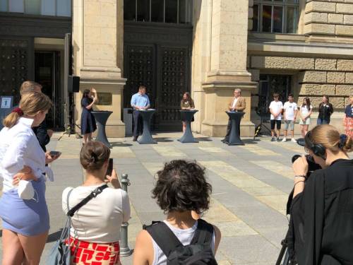 Menschen stehen vor dem Abgeordnetenhaus Berlin im Kreis und hören vier Personen an Stehtischen zu