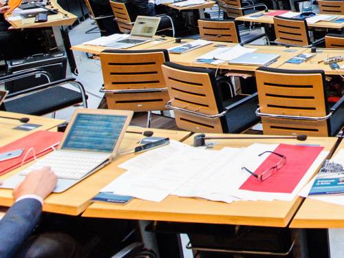 Tische im Plenarsaal. Man sieht eine Hand an einem Laptop und viele Unterlagen.