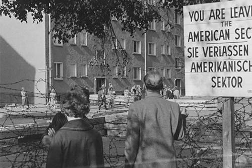 Zwei Menschen blicken am Grenzzaun auf die andere Straßenseite. Neben ihnen ein Schild, auf dem in Englisch und Deutsch steht "Sie verlassen den Amerikanischen Sektor".