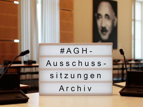 Im Hintergrund ein Sitzungssaal im Anschnitt, im  Vordergrund eine Lightbox mit der Aufschrift "#AGH-Ausschusssitzungen Archiv"