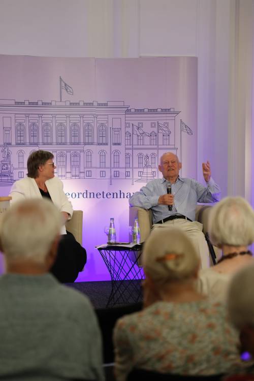 Werner Bork sitzt auf der Bühne in einem Sessel, spricht ins Publikum, hält ein Mikrofon in der einen Hand und gestikuliert mit der anderen.